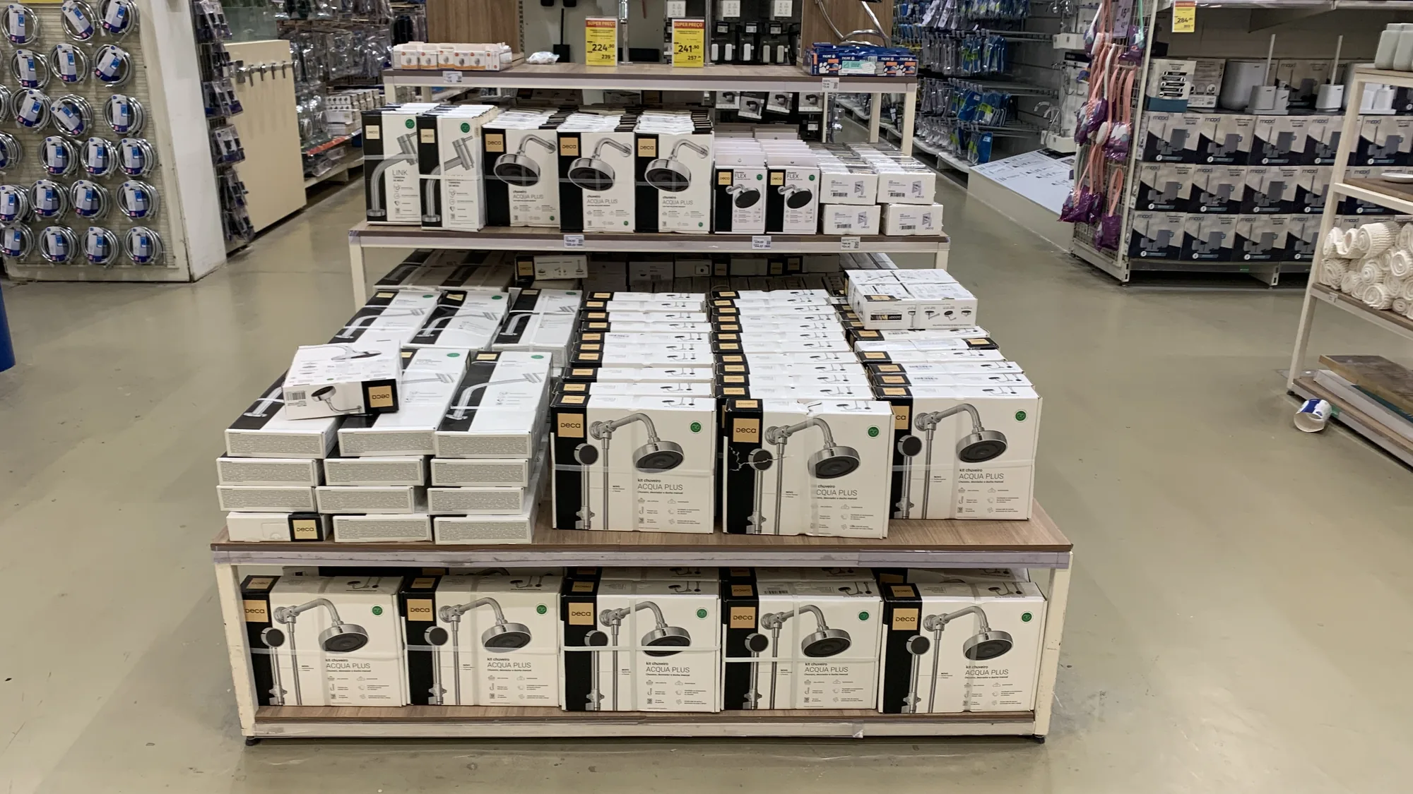 Ilha de promoção de dois níveis no meio do corredor de um home center, com caixas de chuveiro Deca empilhadas e etiquetas de preço.