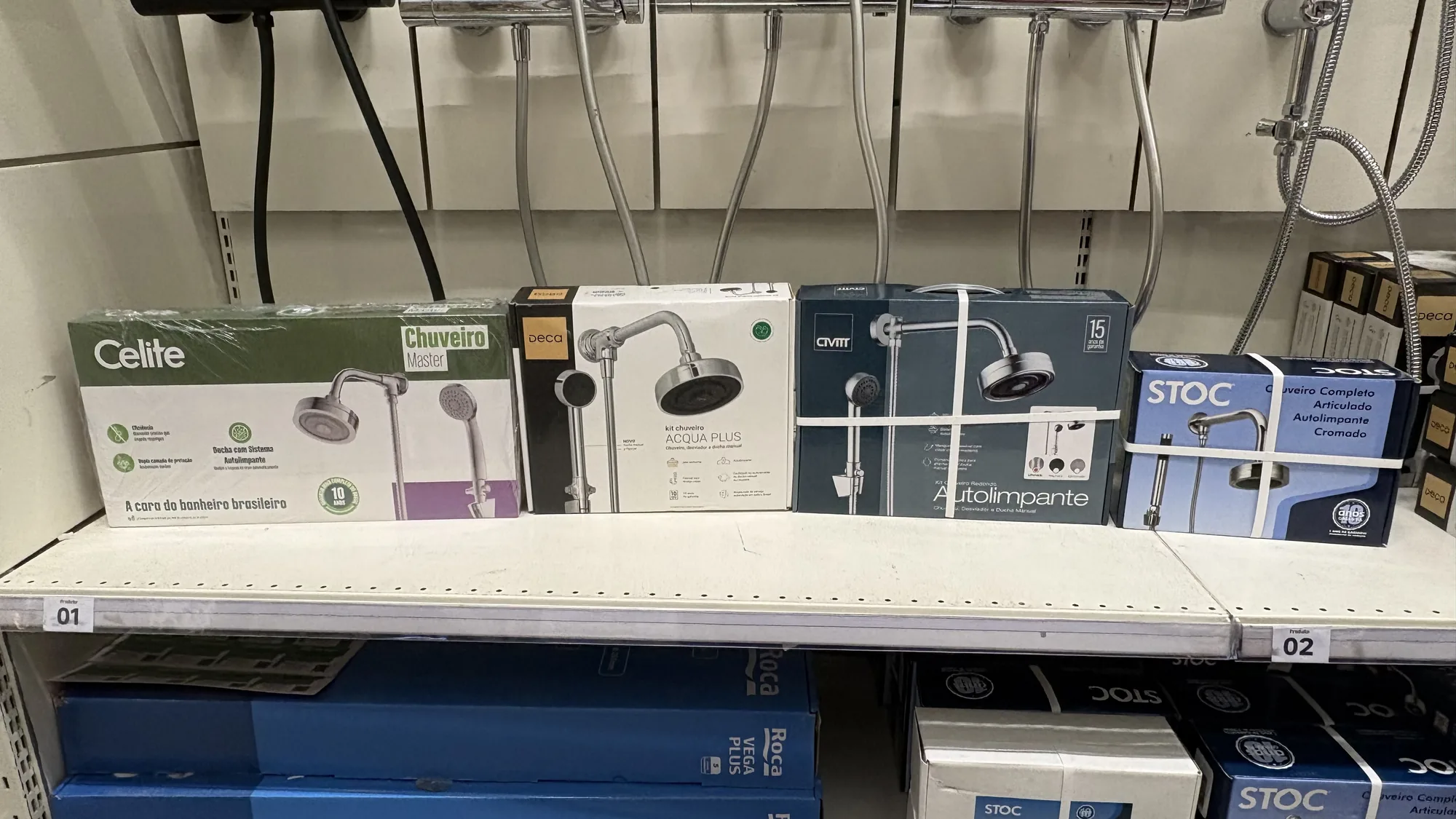 Quatro caixas de chuveiro de marcas diferentes em fila na prateleira de um home center, entre elas o Deca Acqua Plus, com os produtos pendurados acima.
