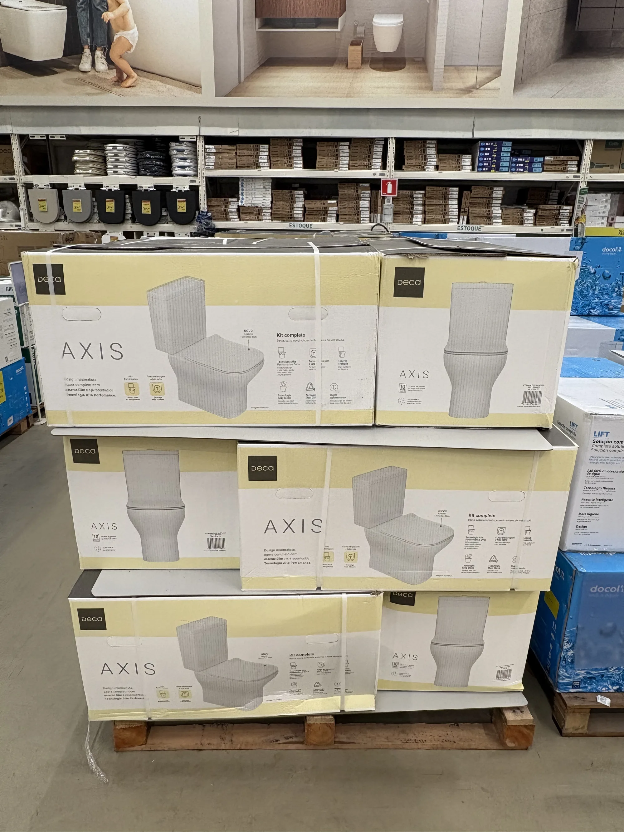Pilha de caixas do vaso sanitário Deca Axis sobre um pallet de madeira, com prateleiras de assentos sanitários ao fundo.