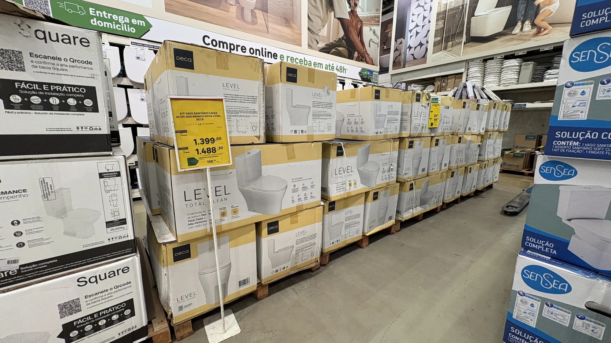 Corredor de home center com vários pallets de caixas do Deca Level alinhados, e expositores da Sensea e da Square nas pontas.