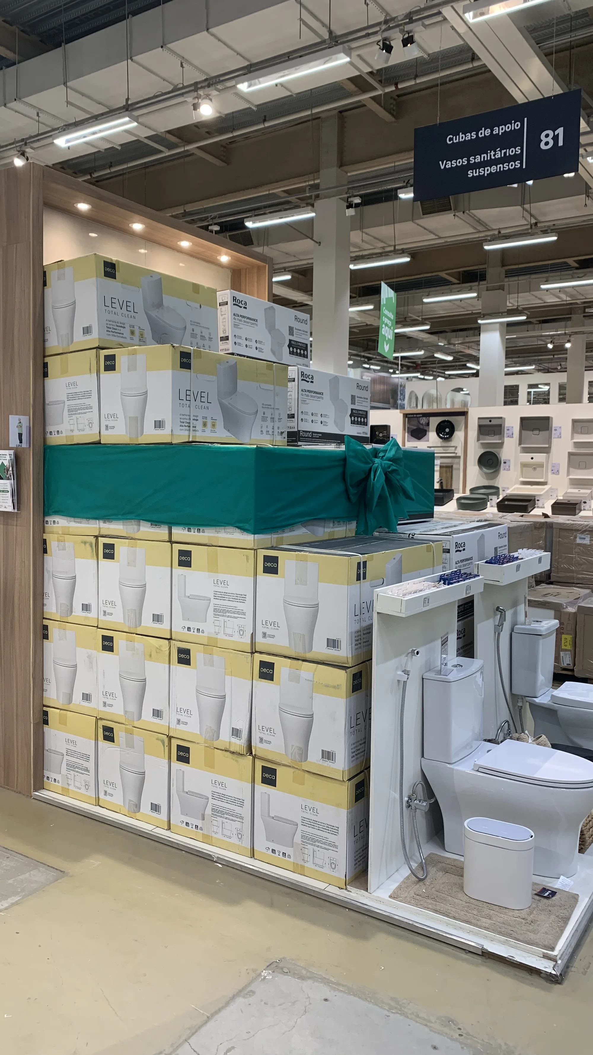 Ponta de gôndola montada com caixas do Deca Level empilhadas até o alto, uma faixa verde com laço no meio e um vaso sanitário exposto ao lado.