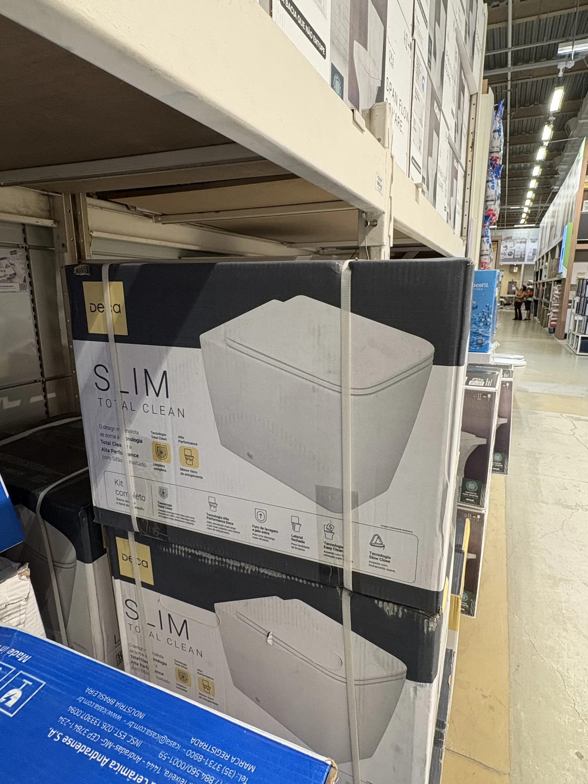Caixas do Deca Slim Total Clean na prateleira alta de um home center, amarradas com fita e vistas de baixo, com o corredor da loja ao fundo.