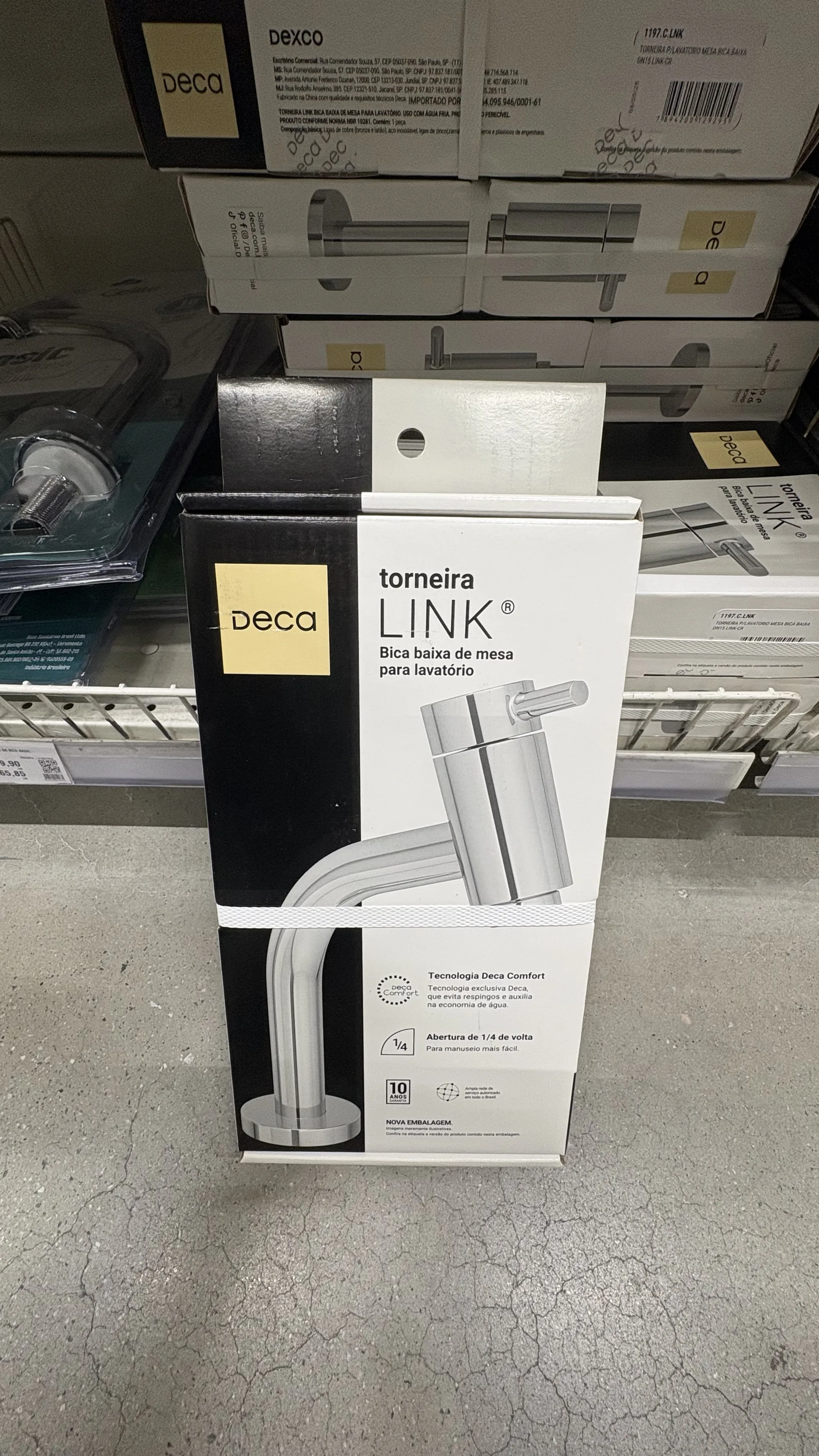 Caixa da torneira Deca Link em pé na prateleira, com o produto impresso na frente em tamanho real e caixas da mesma linha empilhadas ao fundo.