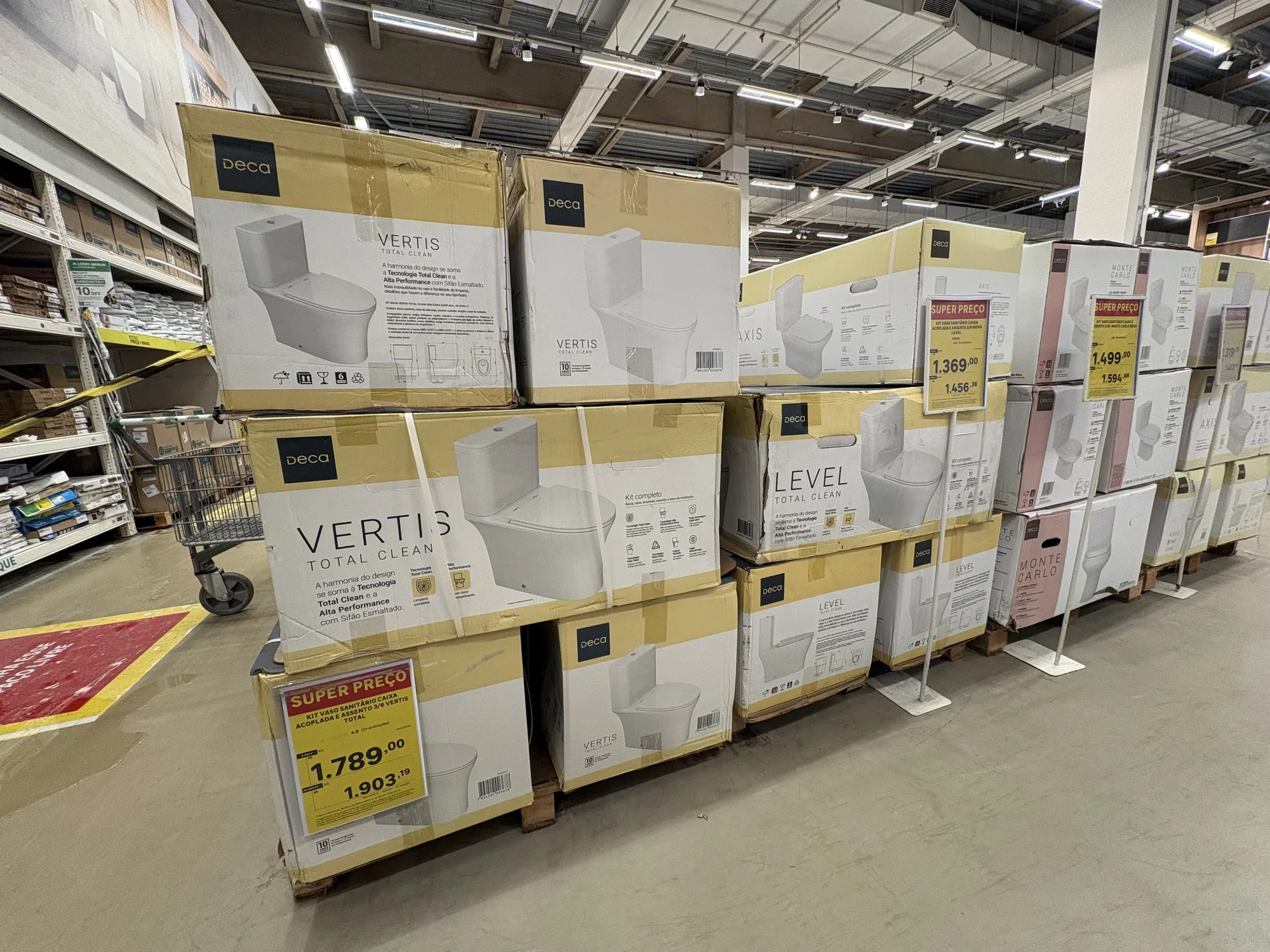 Corredor de louça sanitária com pilhas de caixas Deca Vertis e Deca Level, etiquetas de super preço na frente e sinalização de oferta no chão.