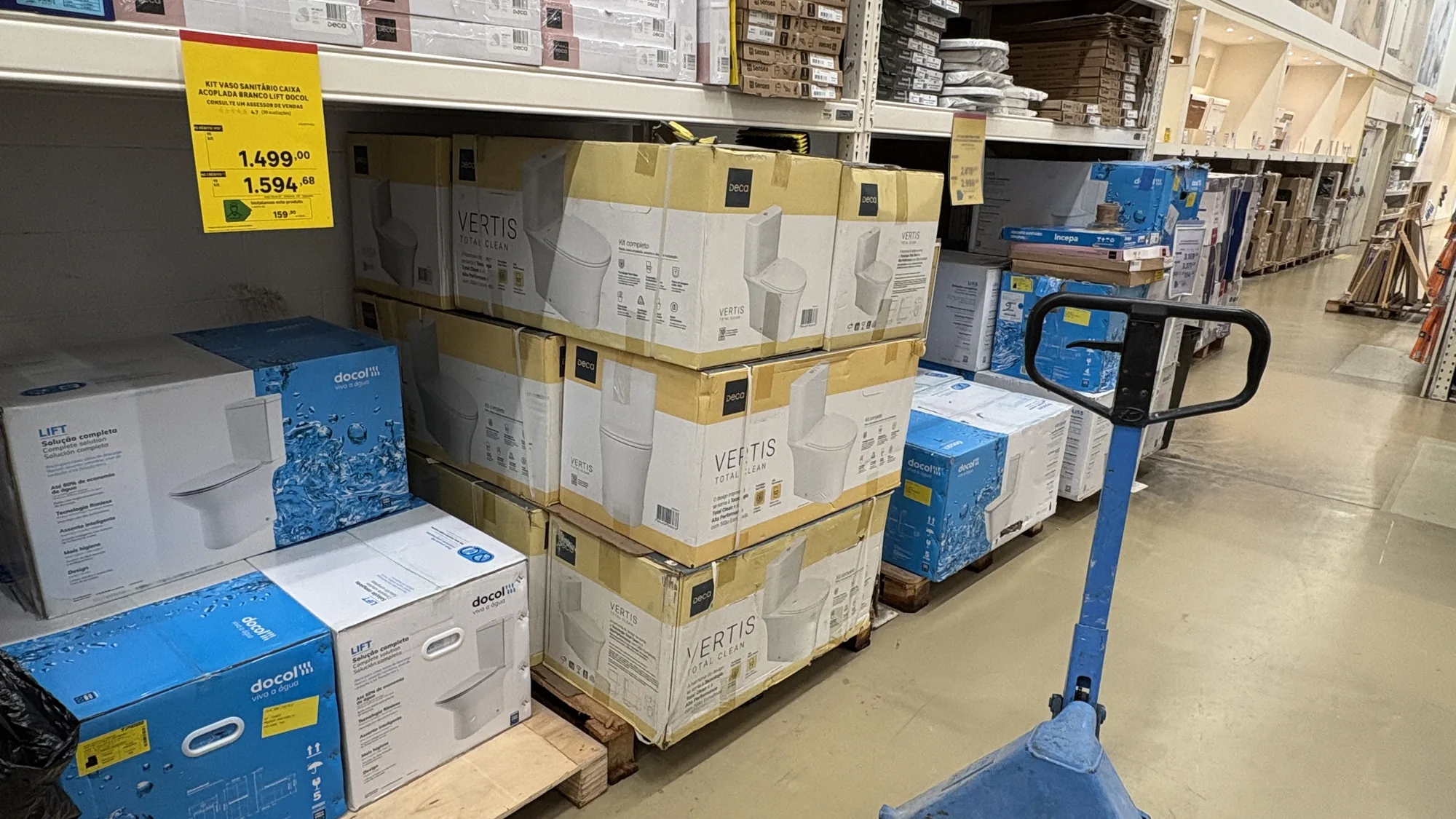 Corredor de um home center com pallets de caixas Deca Vertis, caixas da Docol ao lado e um carrinho hidráulico na frente.