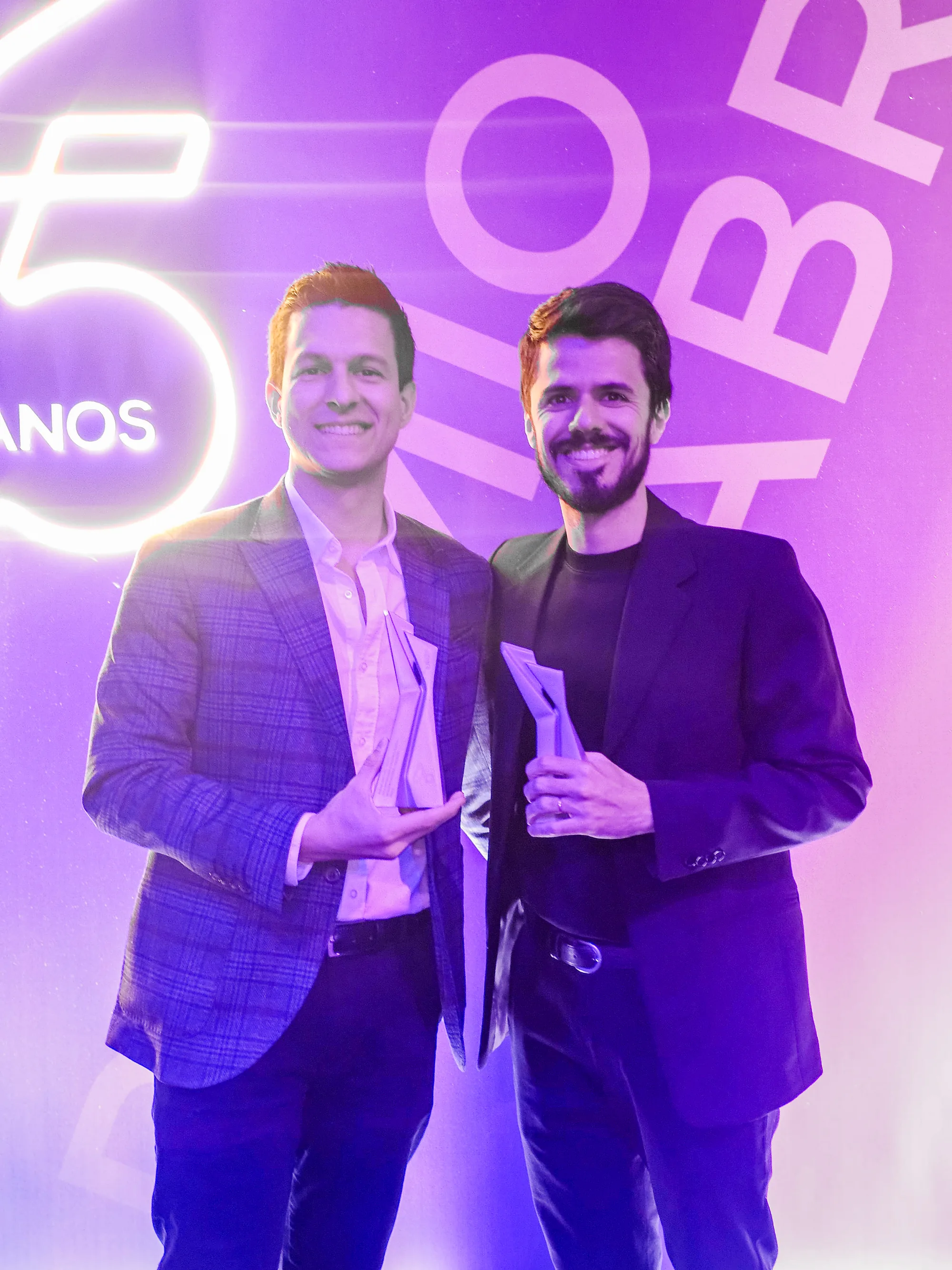 Hugo Zuffo, on the left, and one other person holding the silver trophies of the Prêmio ABRE da Embalagem Brasileira 2025, in front of the lit backdrop of the ceremony.
