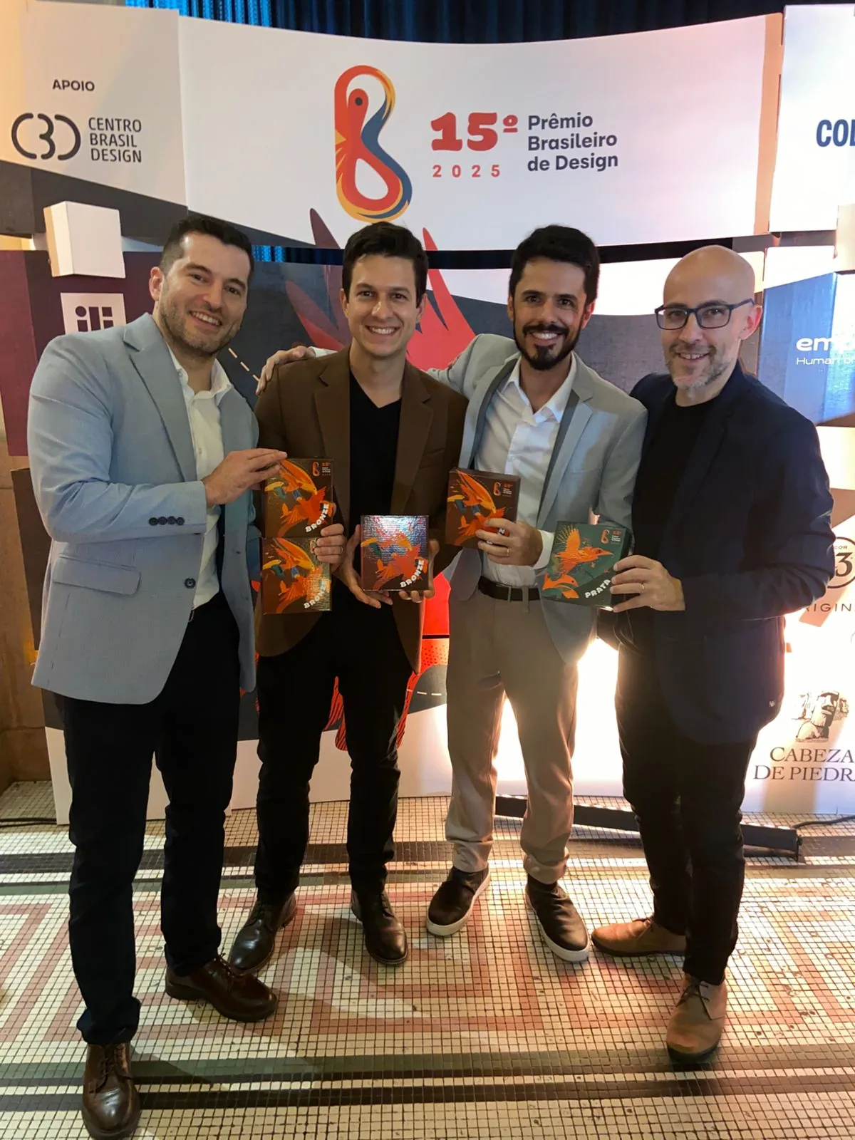 Hugo Zuffo, second from the left, with three other people holding the trophies of the 15th Brazilian Design Award, in front of the event backdrop.